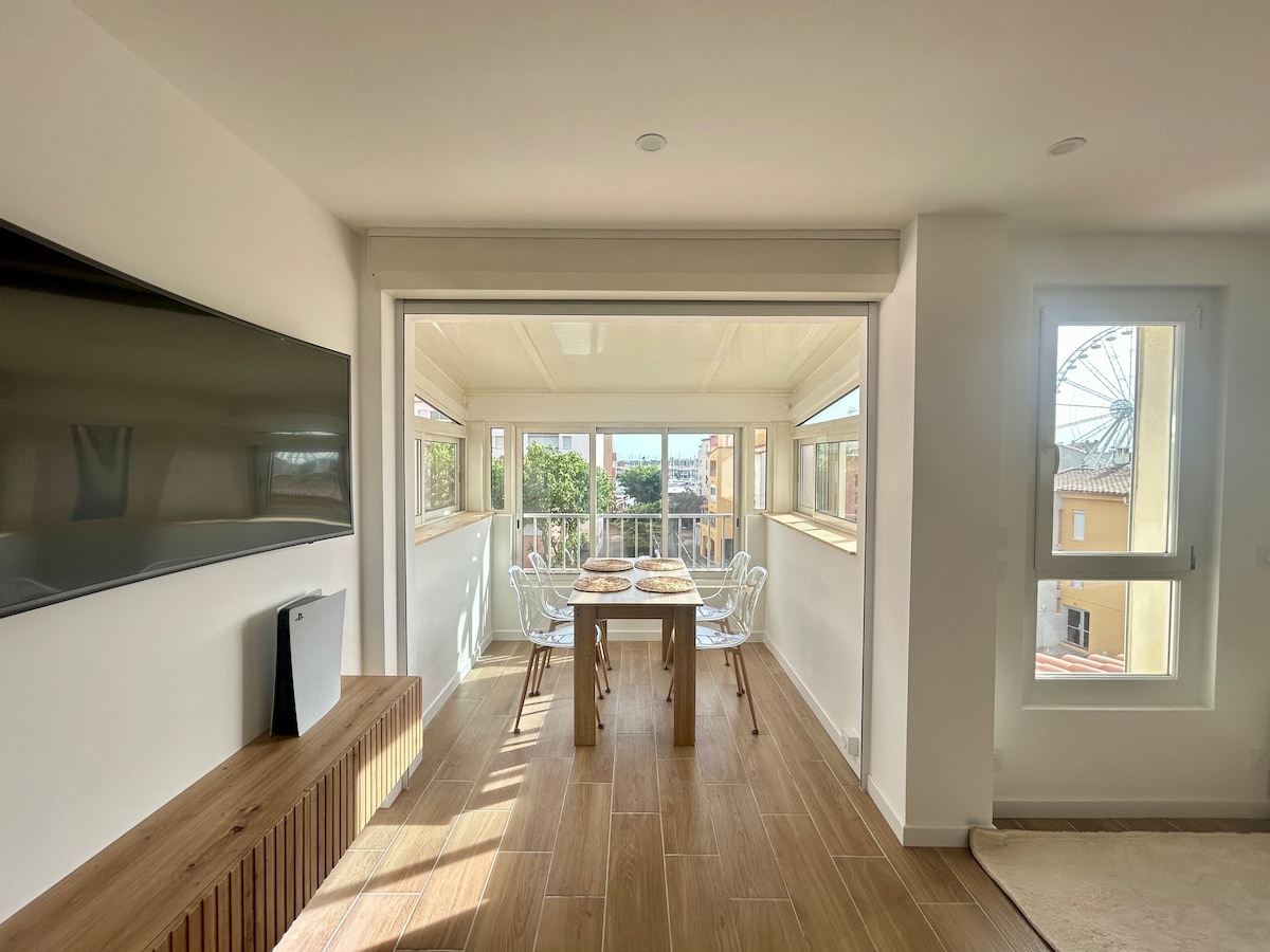 A bright living space features large windows offering views of the surroundings. A wooden dining table is set with tableware, surrounded by transparent chairs. A modern television is mounted on the white wall, complemented by a wooden console below, enhancing the room's welcoming ambiance.