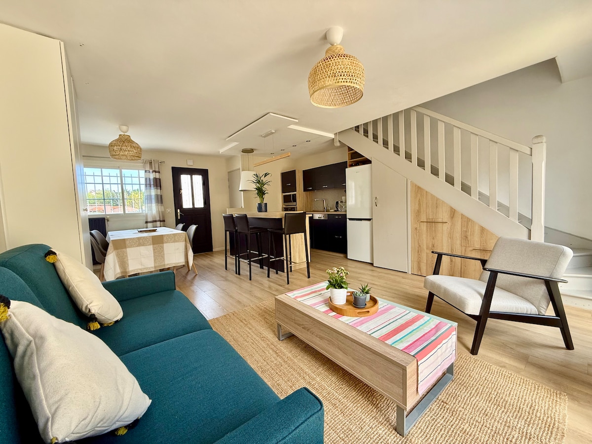 A bright living area features a comfortable sofa and a stylish armchair. Natural light enters through large windows, illuminating a dining table and a fully equipped kitchen in the background. A woven rug adds texture to the space, creating a cozy atmosphere.