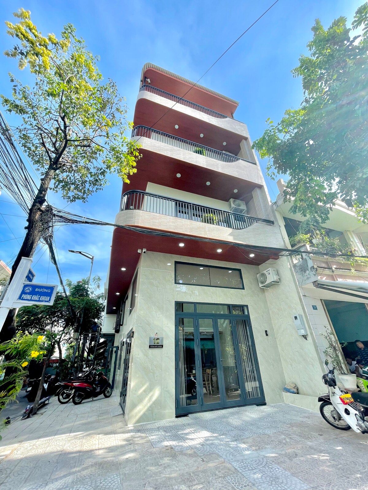 A three-story building is visible, featuring a modern exterior with large glass doors and multiple balconies. Sunlight illuminates the facade, highlighting the clean lines and contemporary design. Surrounding greenery adds a touch of nature to the urban setting.