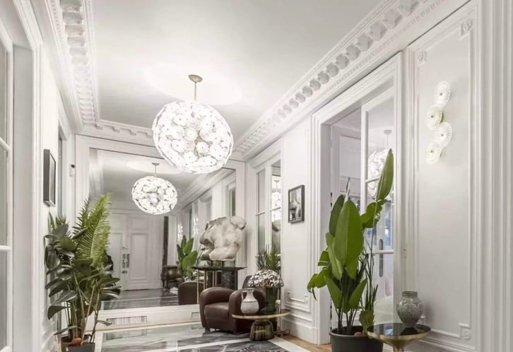 A bright hallway is highlighted by reflective surfaces and artistic light fixtures. Lush green plants are positioned along the sides, complementing the elegant decor. A sculpture and framed artwork are displayed against the walls, enhancing the space's sophistication.