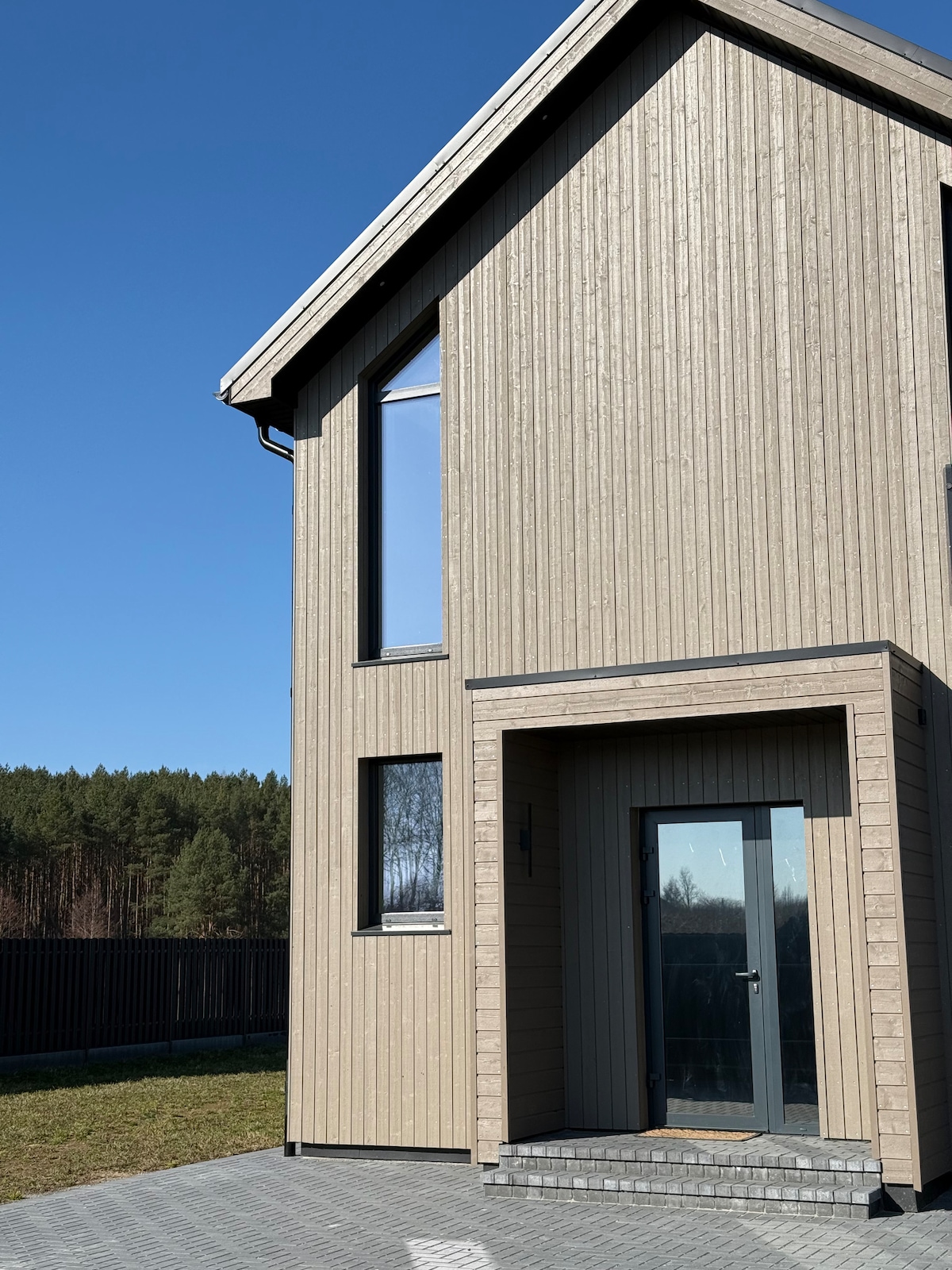 A modern house exterior is shown, featuring a light wood panel facade and large windows that allow natural light. The entrance is framed by a stone step leading to a sleek front door. A clear blue sky and green trees form the backdrop.