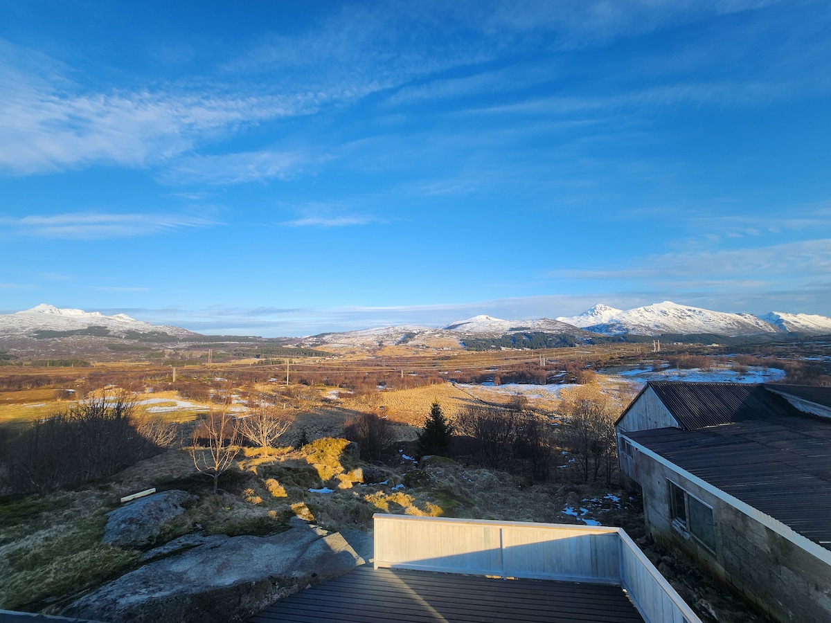 A panoramic view captures a vast landscape with rolling hills and snow-capped mountains in the distance. Clear blue skies dominate, while patches of green and brown fields are visible, indicating the changing seasons. A quaint, older structure sits in the foreground.