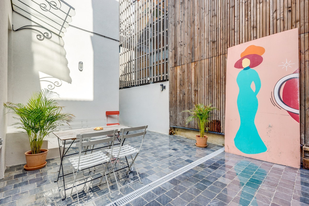A private terrace features a tiled floor and is furnished with a table and chairs for outdoor dining. Potted plants add greenery, while a colorful mural of a woman enhances the space's character. A wooden wall provides privacy, creating a cozy ambiance.