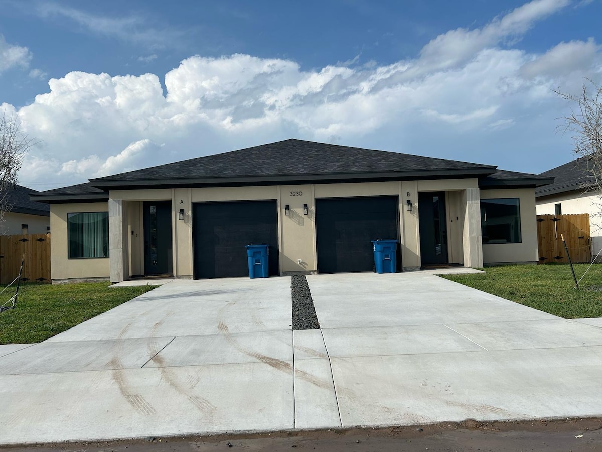 The exterior of a newly built duplex is presented, showcasing a symmetrical design with two dark garage doors. A concrete driveway leads to the entrance, flanked by green grass and a wooden fence. Blue trash bins add a touch of color against the neutral tones of the structure.