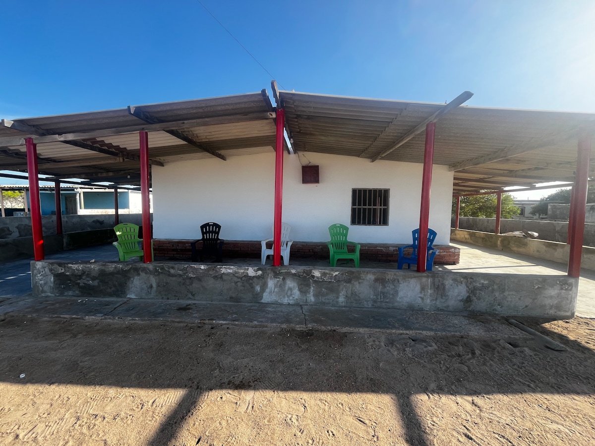 A covered outdoor area is shown, featuring a row of colorful plastic chairs in green, blue, and black. The white walls of the structure are contrasted by red support beams, with sunlight creating clear shadows on the ground. The surrounding sandy area adds to the setting.