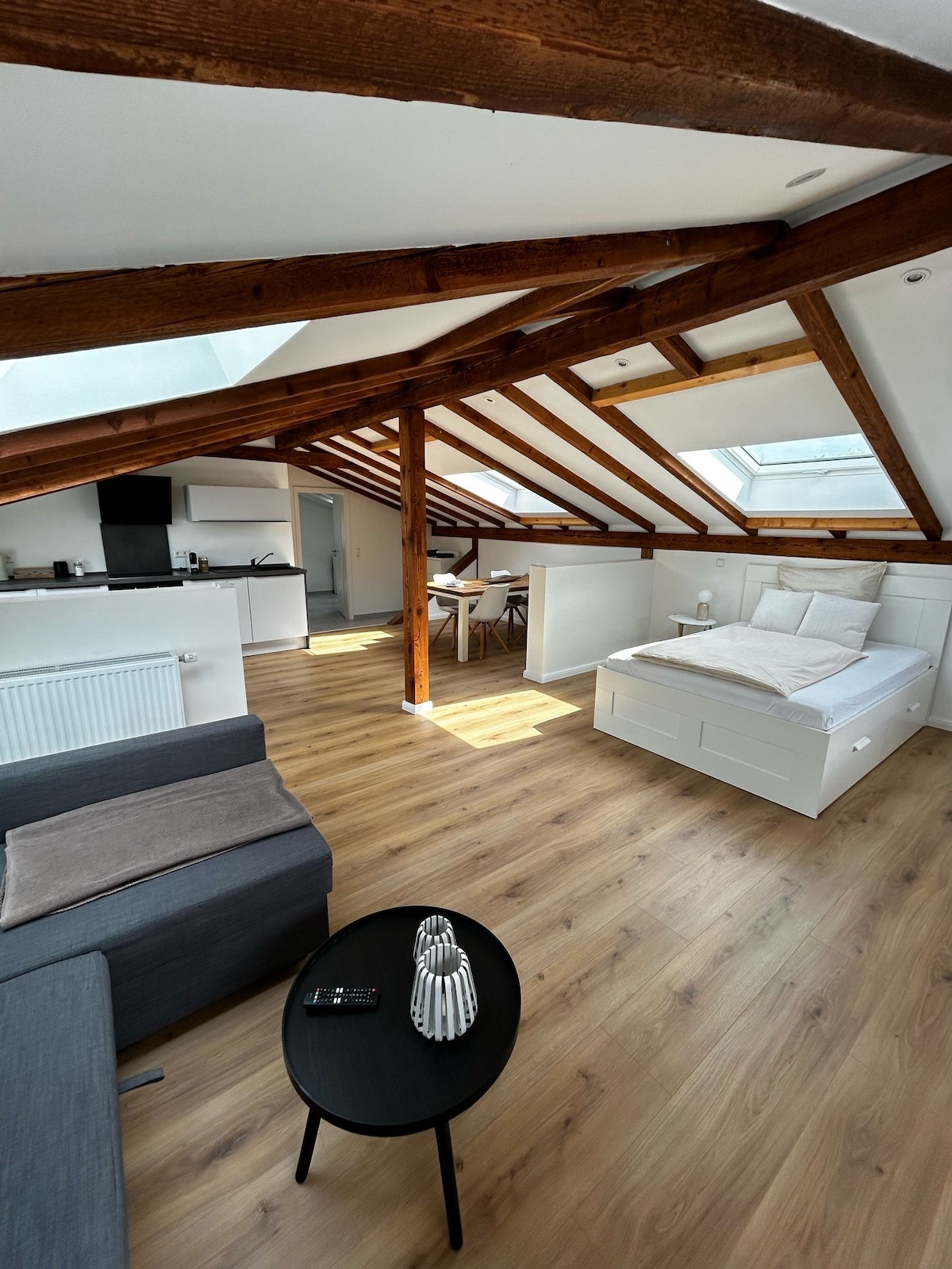 An open living space features exposed wooden beams and natural light from skylights. A spacious bed is positioned adjacent to a modern sofa and a small black table. A dining area is visible in the background, complementing the minimalistic decor.