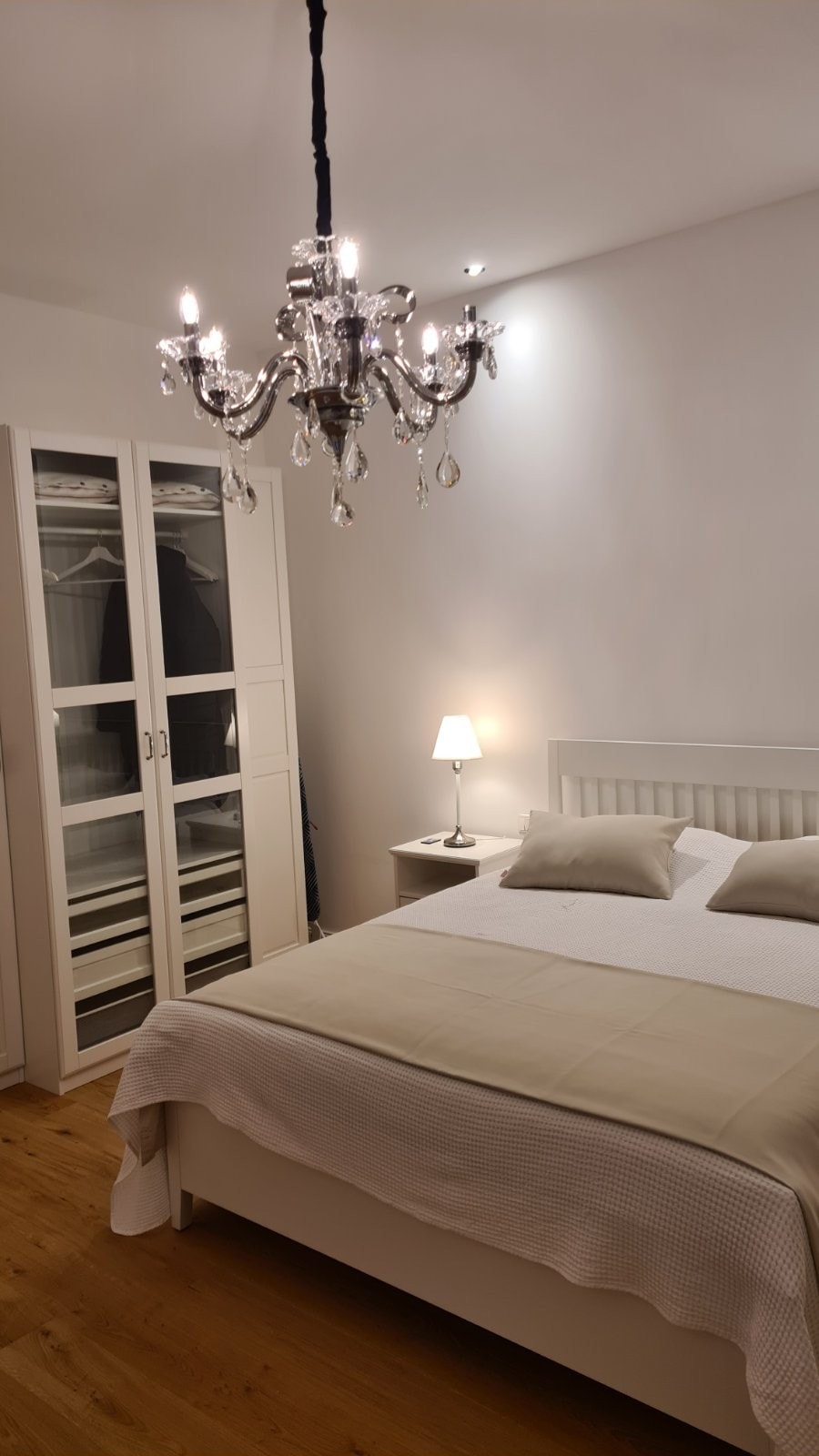 A well-lit bedroom showcases a spacious bed with neutral-toned linens, complemented by decorative pillows. A stylish chandelier hangs from the ceiling, while a compact nightstand with a lamp is placed beside the bed. A wardrobe with glass doors offers additional storage options.