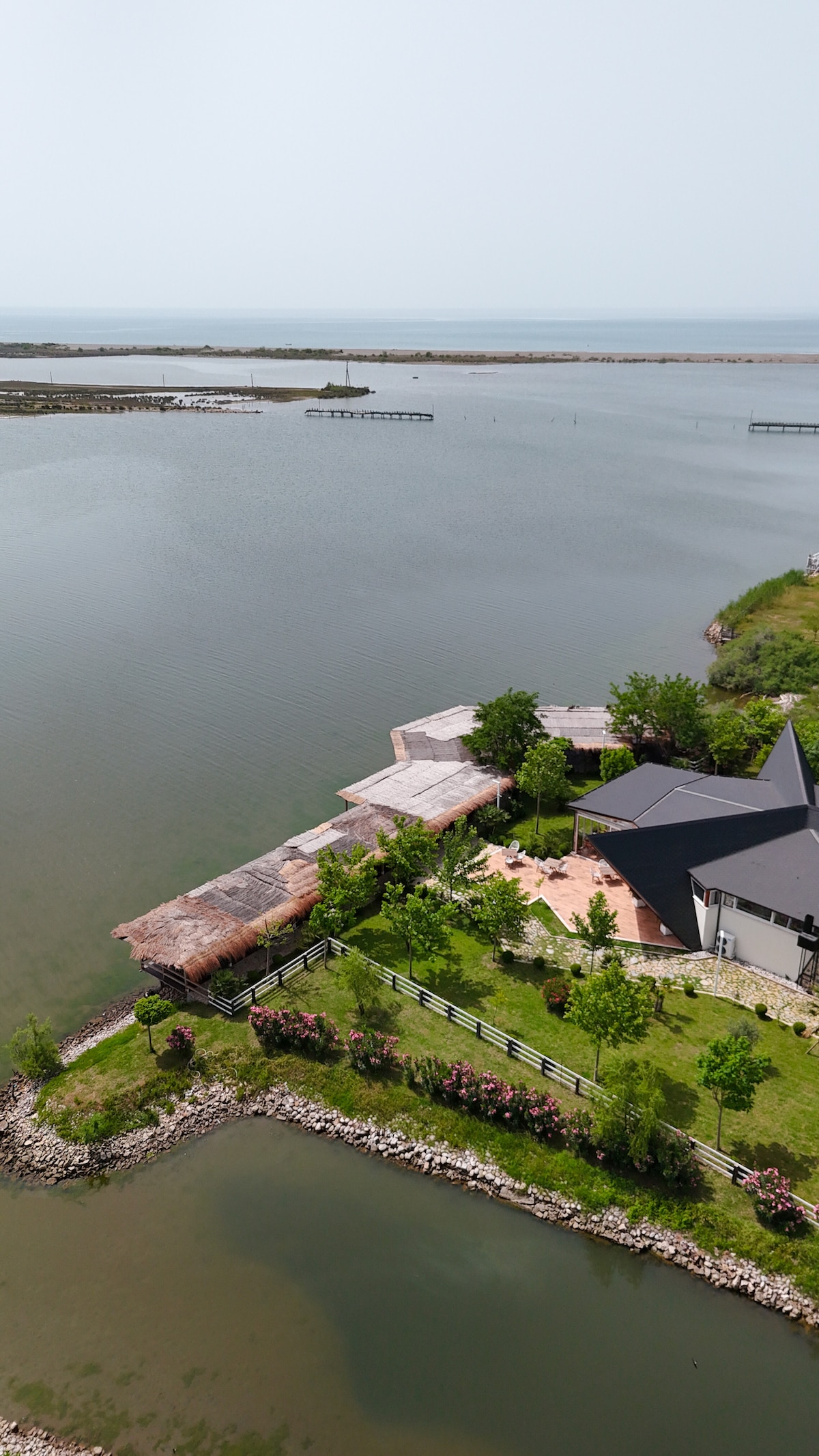 A serene aerial view captures the campsite nestled beside a calm body of water. Lush greenery surrounds the structures, and a wooden pier extends into the water. The gentle curves of the landscaping lead the eye towards distant views of the open sea.