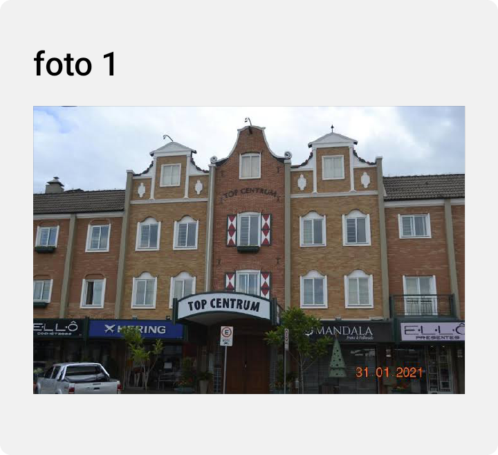 The image shows a multi-story building featuring a symmetrical facade with a prominent sign displaying 'TOP CENTRUM.' Windows are framed in white, and the entrance is marked by an awning. Landscaping elements, including plants, are visible at the base of the structure.