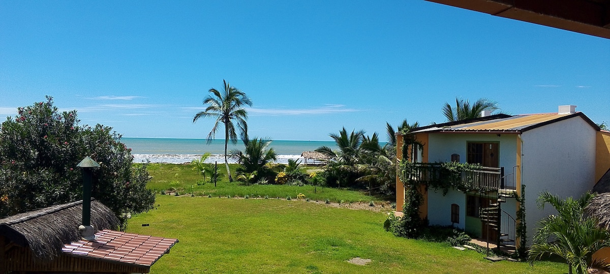 A lush green yard featuring palm trees and shrubs leads to a sandy beach visible in the background. Two buildings are positioned within the garden, one of which displays a balcony. The bright blue sky complements the serene beachfront setting.