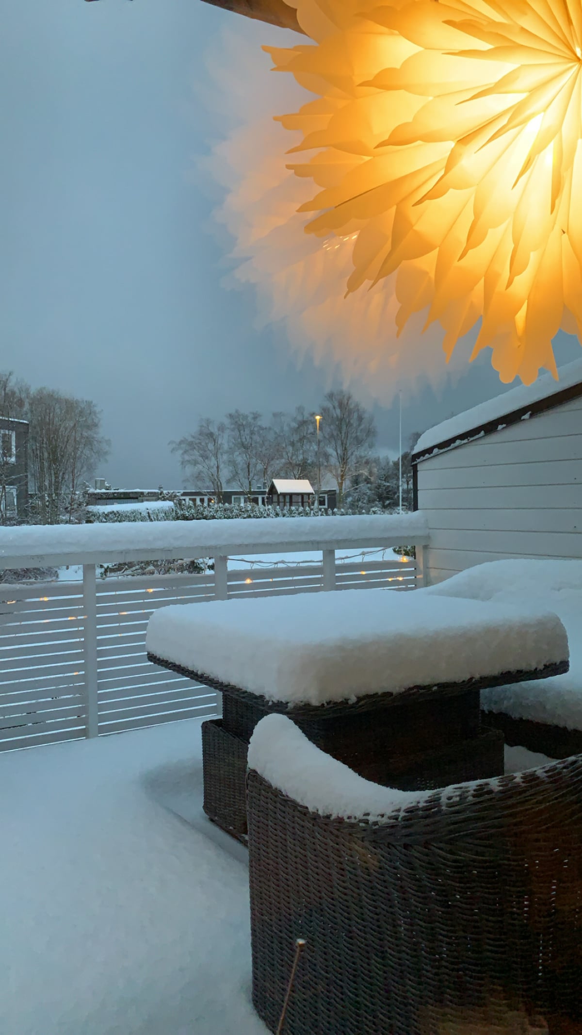 A winter scene is depicted on a private balcony covered in fresh snow. A large decorative light fixture hangs above a table and seating area, providing a warm glow against the cool outdoor landscape. Snow blankets the furniture, creating a serene atmosphere.