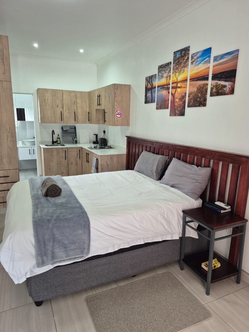 A cozy bedroom is presented with a spacious bed dressed in white linens, flanked by two pillows. A small side table holds a lamp and personal items. Light wooden cabinetry and kitchen appliances are visible in the background, while framed nature photographs adorn the wall.