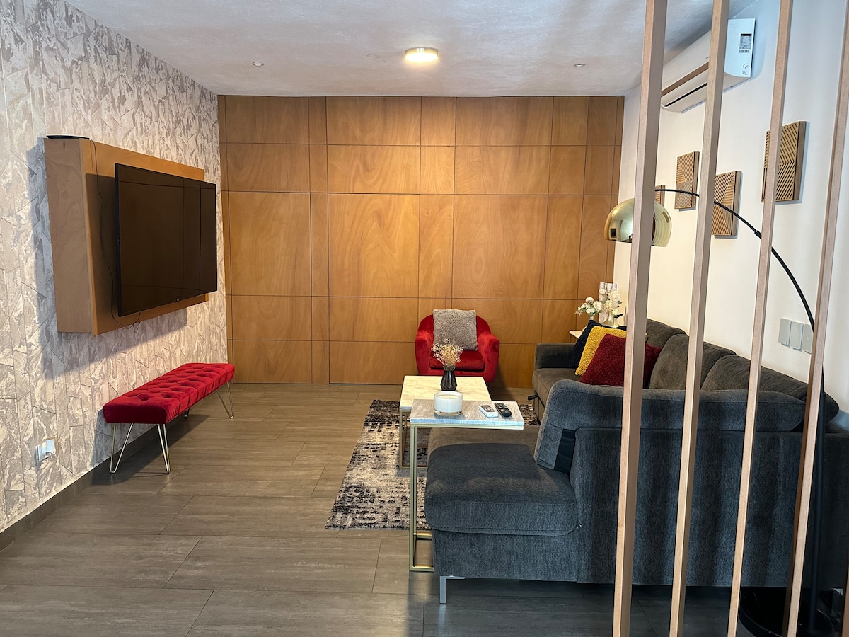 A modern living area features a dark sofa complemented by red armchairs. A wooden wall hosts a flat-screen television, while a sleek white coffee table rests on a patterned rug. Ambient lighting casts a warm glow over the space, enhancing the overall design.