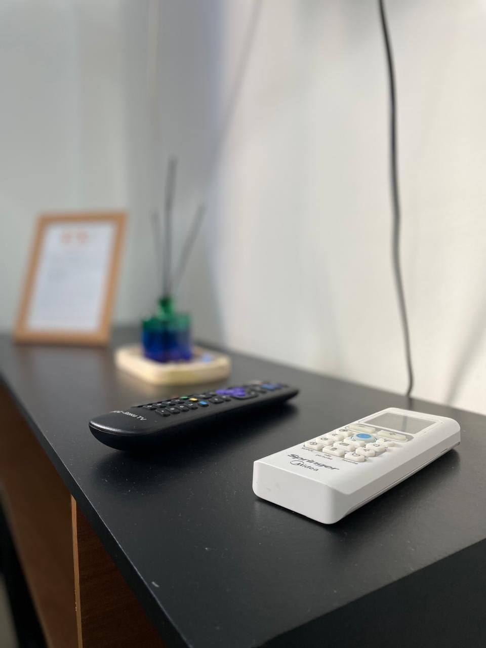 A sleek black tabletop displays a remote control and a white device, possibly for the Smart TV. A decorative item, possibly a vase with sticks, adds a touch of color, while a framed informational card rests nearby, enhancing the organized environment.