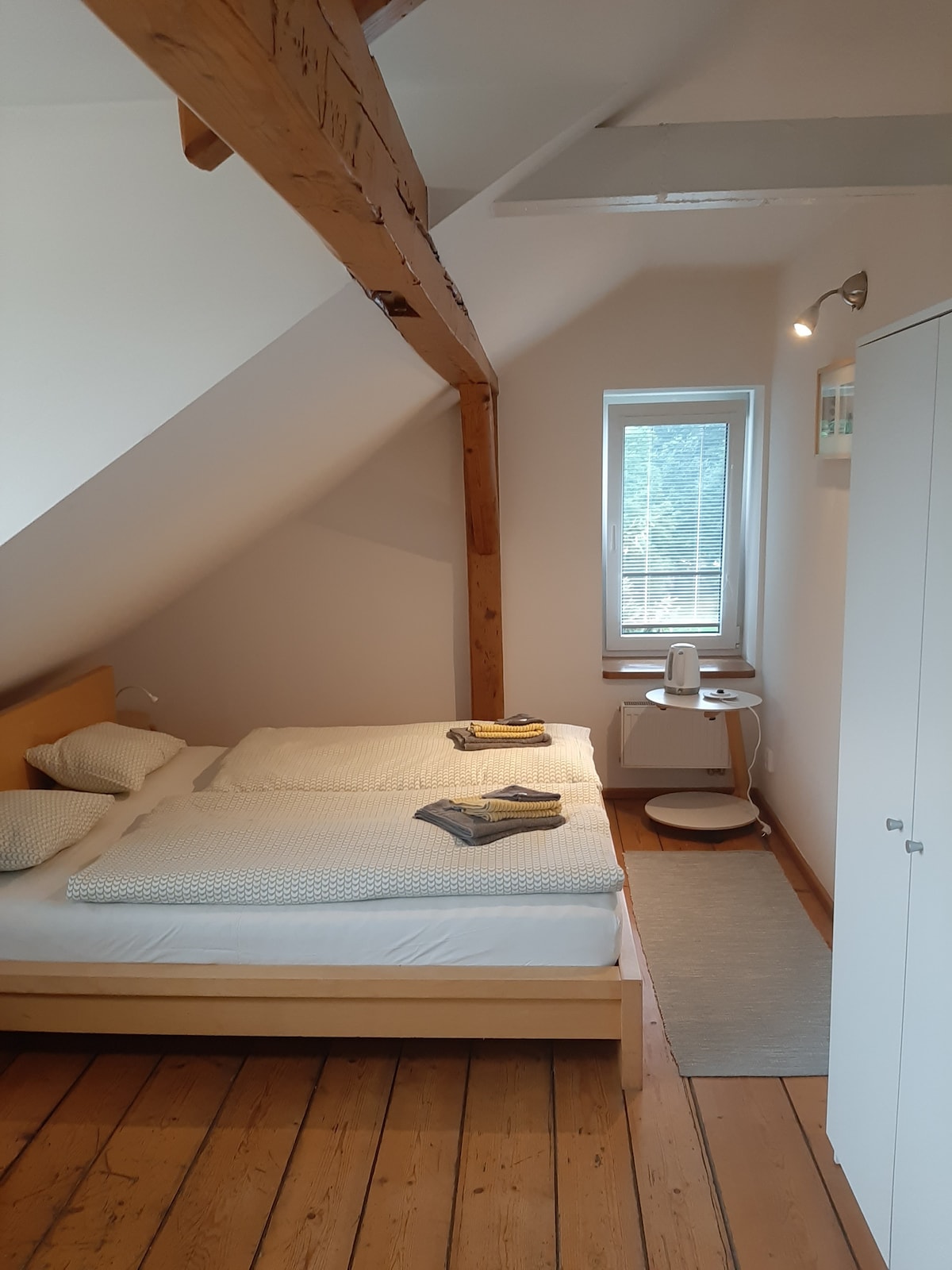 A cozy sleeping area is defined by wooden beams and slanted ceilings. Two spacious beds are positioned against a light-colored wall, adorned with neatly arranged linens. A small nightstand and a chair are located nearby, with a window allowing natural light to enter.