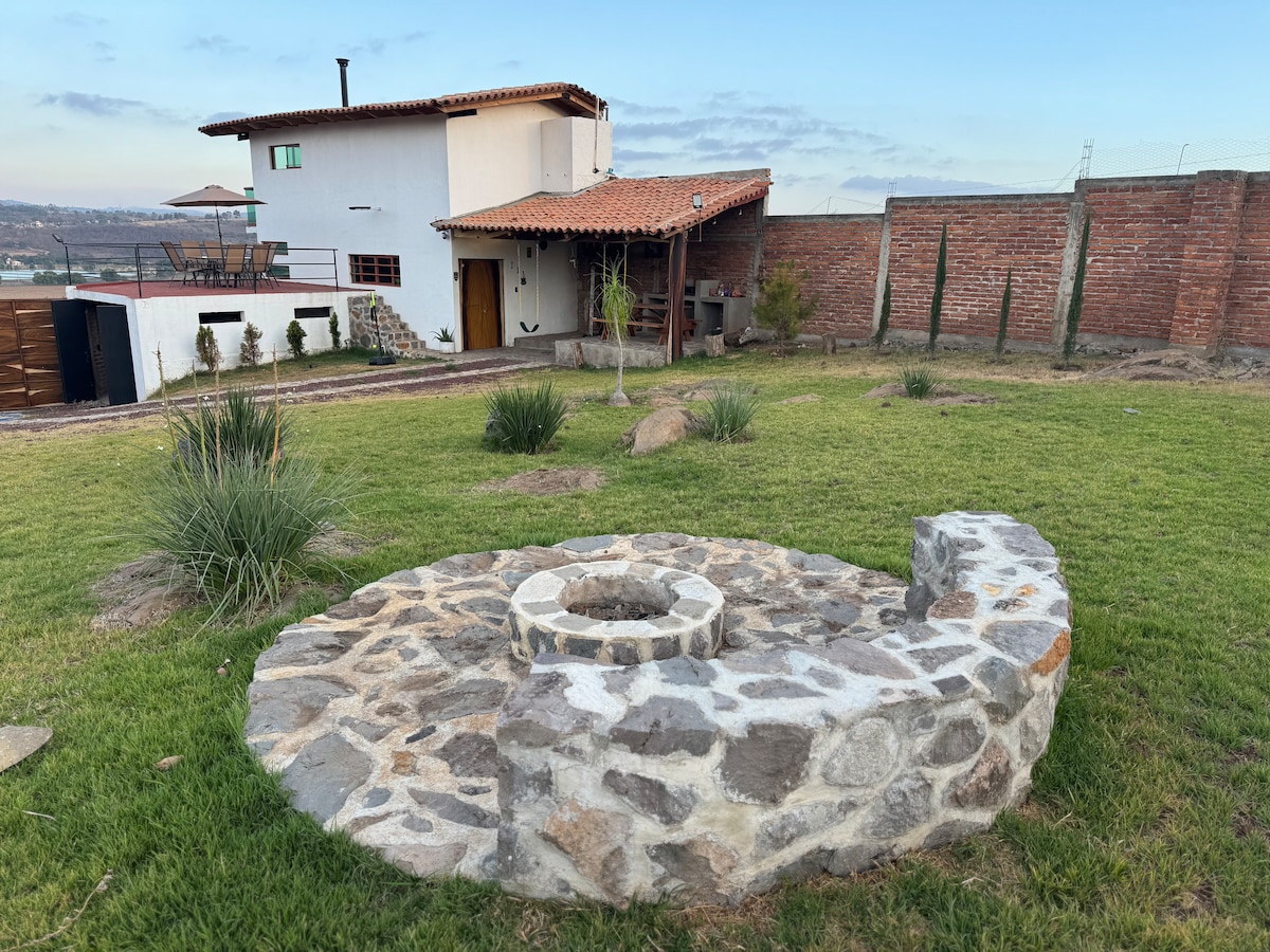 A circular stone fire pit is situated on a grassy area, surrounded by sparse landscaping. In the background, a two-story white building with a rustic, tiled roof is visible, along with a wooden structure to the side. A clear blue sky complements the serene outdoor setting.