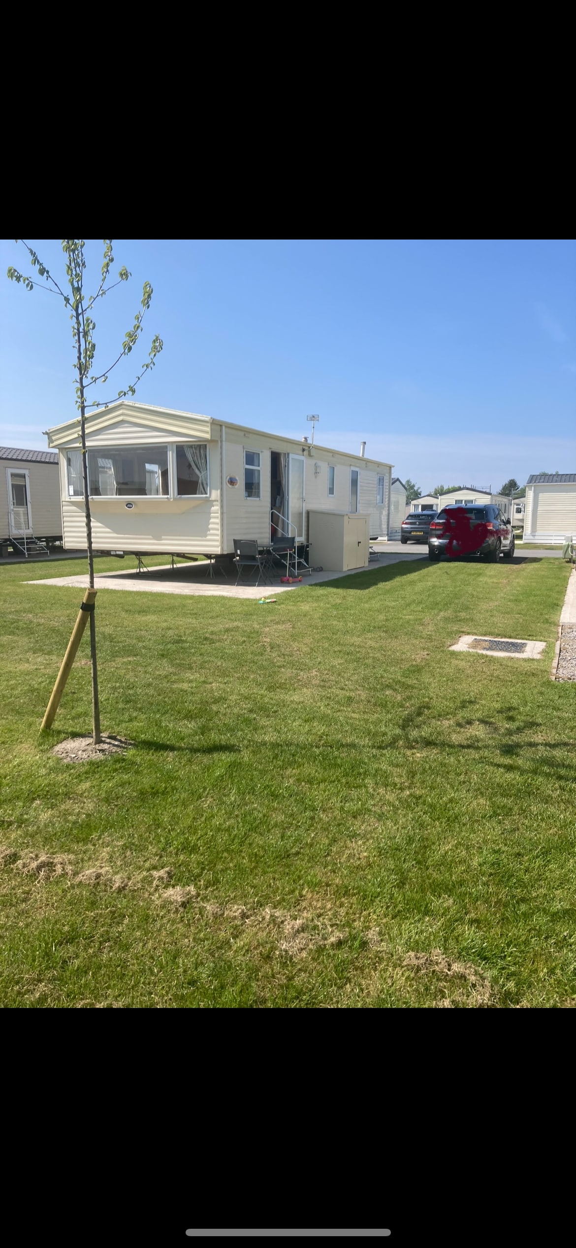 A static caravan is set on a neatly maintained grassy area, accompanied by a small tree. The exterior of the caravan features light yellow siding and steps leading to the entrance. A vehicle is parked nearby, while neighboring caravans are visible in the background.
