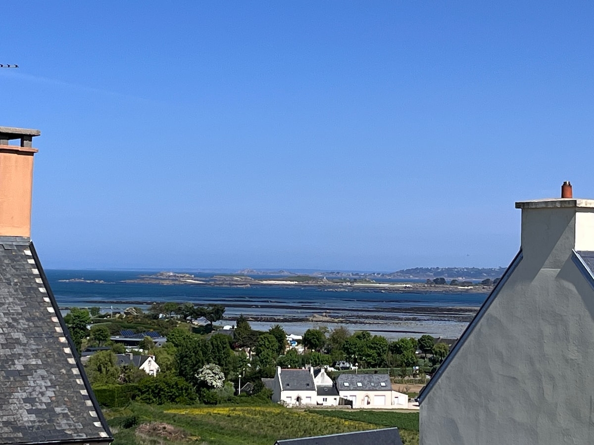 A scenic view showcases the coastline and distant islands, framed by the rooftops of nearby houses. The clear blue sky and tranquil sea complement the greenery of the landscape, offering a sense of openness and connection to nature.