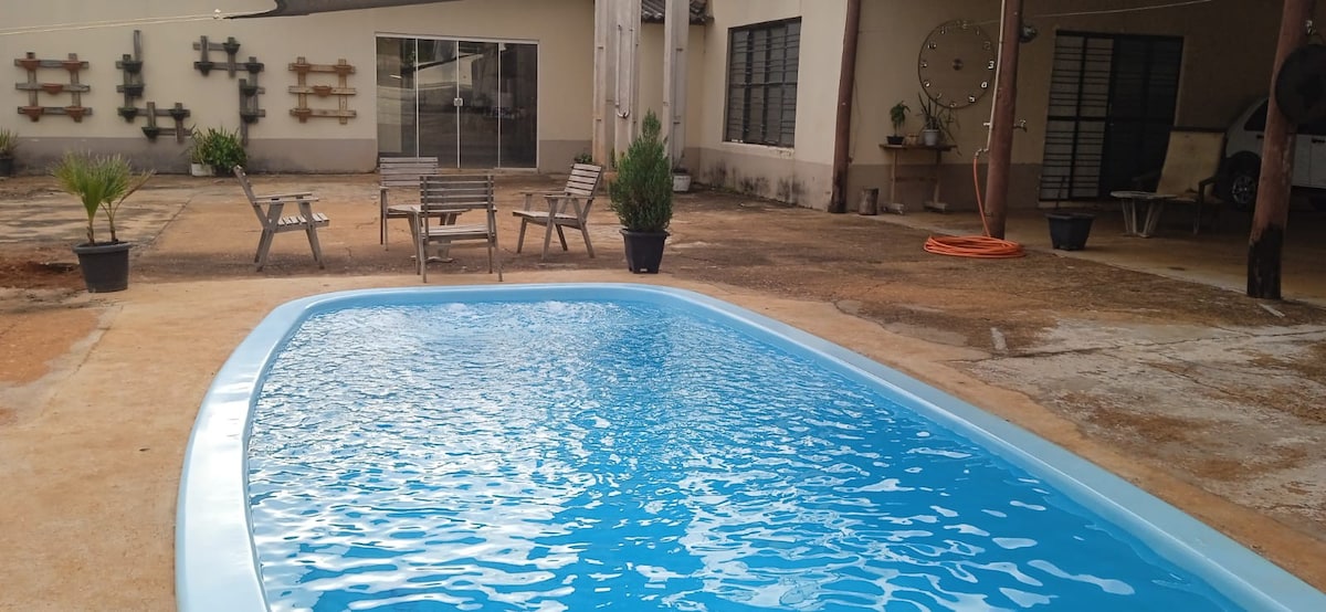 A clear swimming pool occupies the foreground, surrounded by a spacious outdoor area. Several lounge chairs are arranged nearby, alongside potted plants. In the background, large glass doors lead to a building, enhancing the open atmosphere.