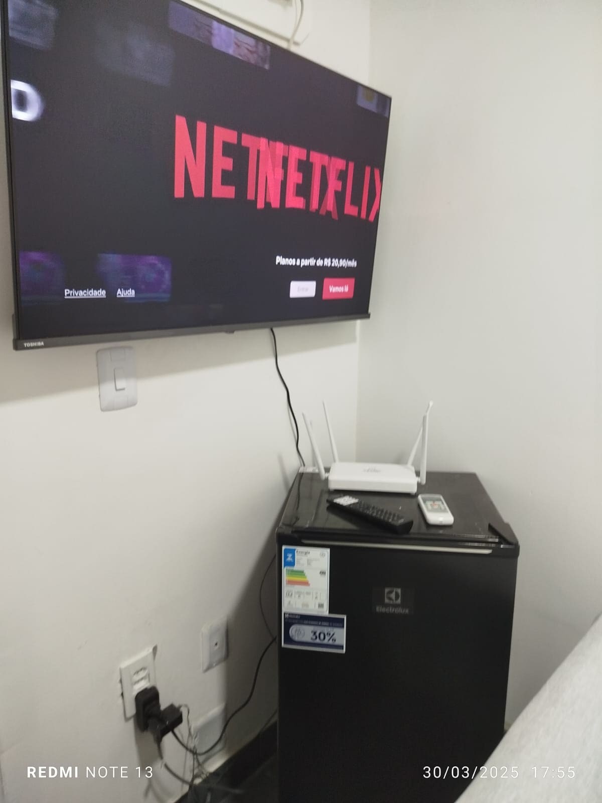A compact entertainment area features a Smart TV displaying a streaming service logo above a small refrigerator. A Wi-Fi router is positioned beside the fridge, which includes a visible energy efficiency label. The setting provides essential amenities for a comfortable stay.