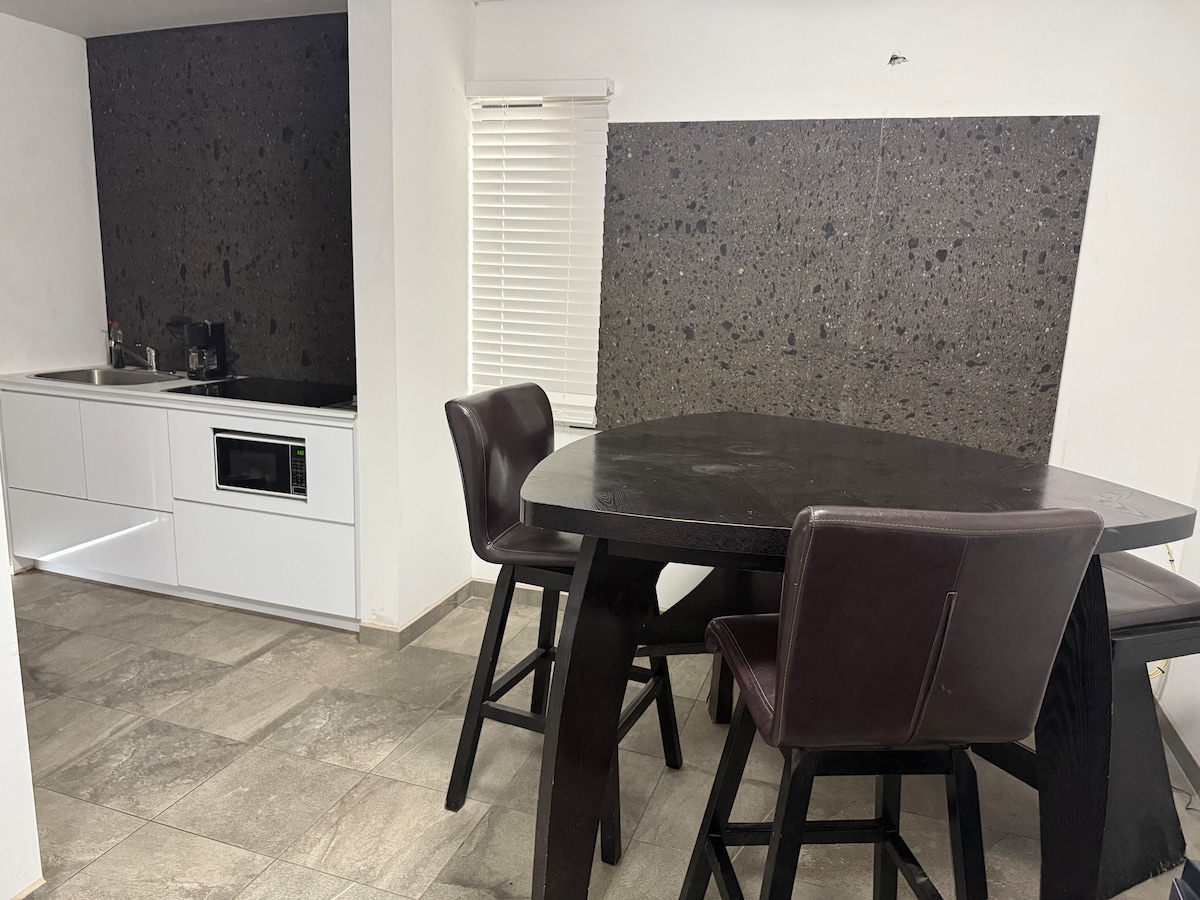 A dining area features a round black table with four dark chairs. A minimalist kitchen is visible in the background, showcasing sleek white cabinetry and a microwave. A textured, dark stone wall serves as a striking backdrop for the space.