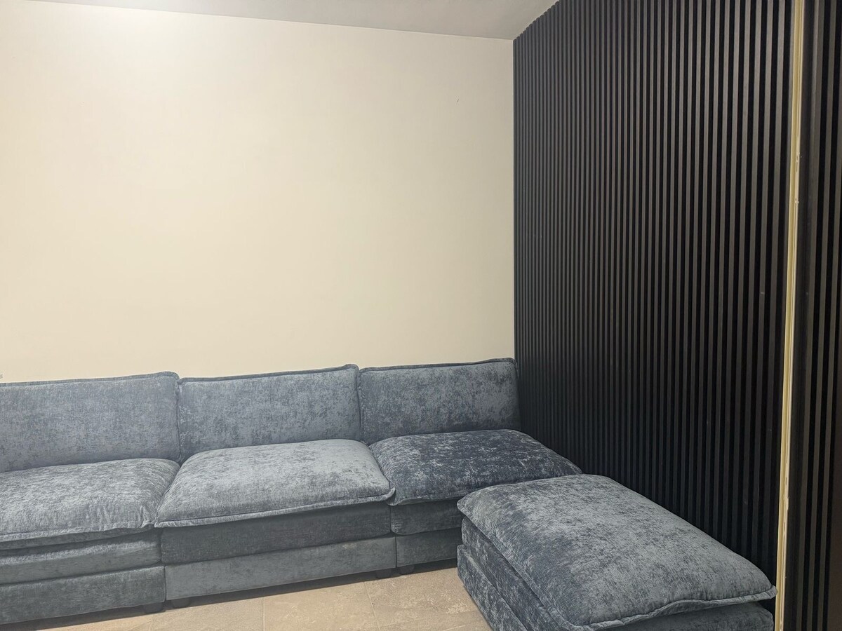 A spacious living area is furnished with a large sectional sofa, upholstered in soft blue fabric. The wall is adorned with vertical black paneling, contrasting with the light-colored wall opposite. A coordinated ottoman complements the seating arrangement, providing additional comfort.