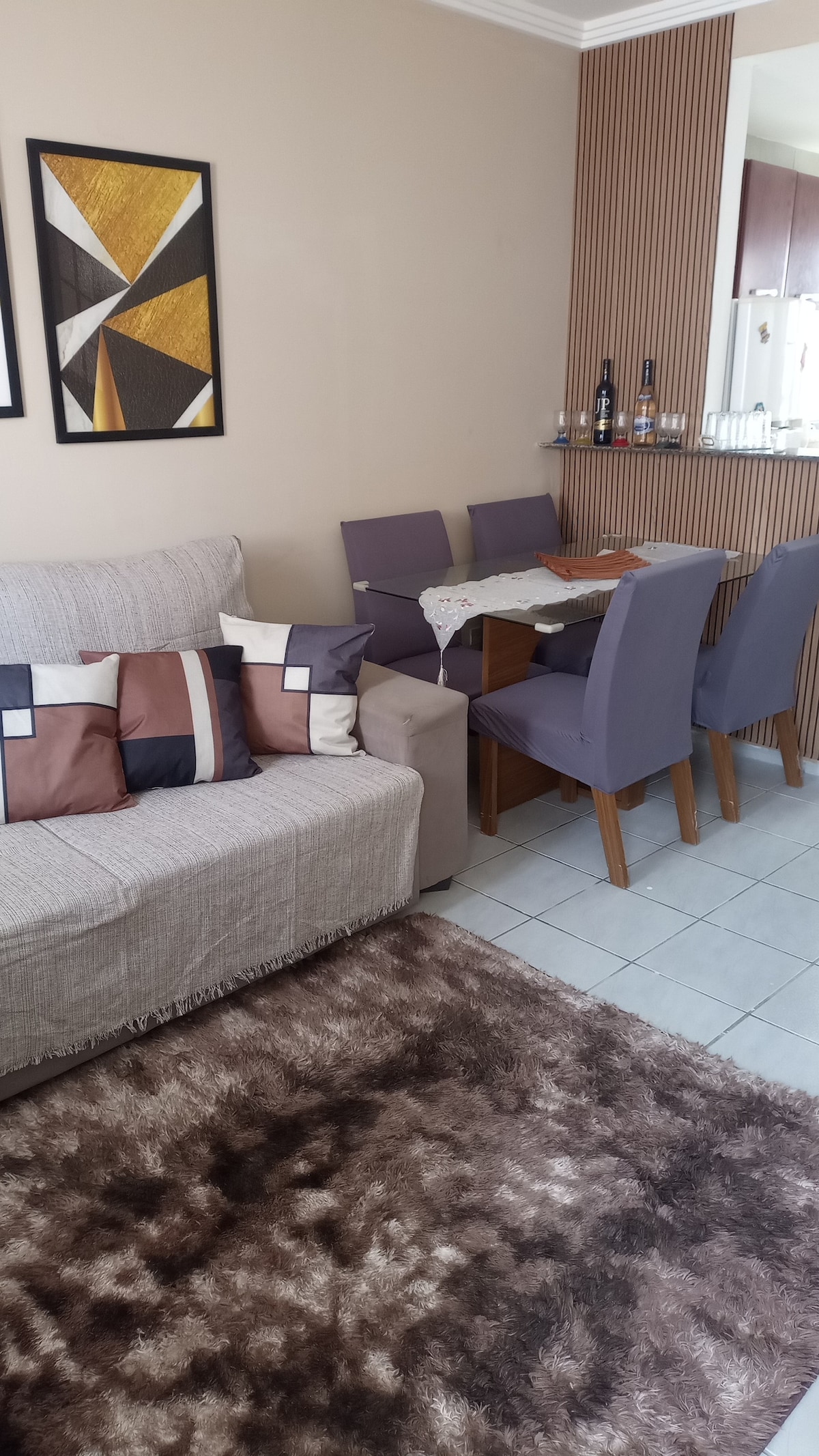 A cozy living space features a soft sofa adorned with a mix of patterned cushions. A small dining area with a neatly set table and four upholstered chairs is present. The floor is covered by a plush rug, adding texture to the room.