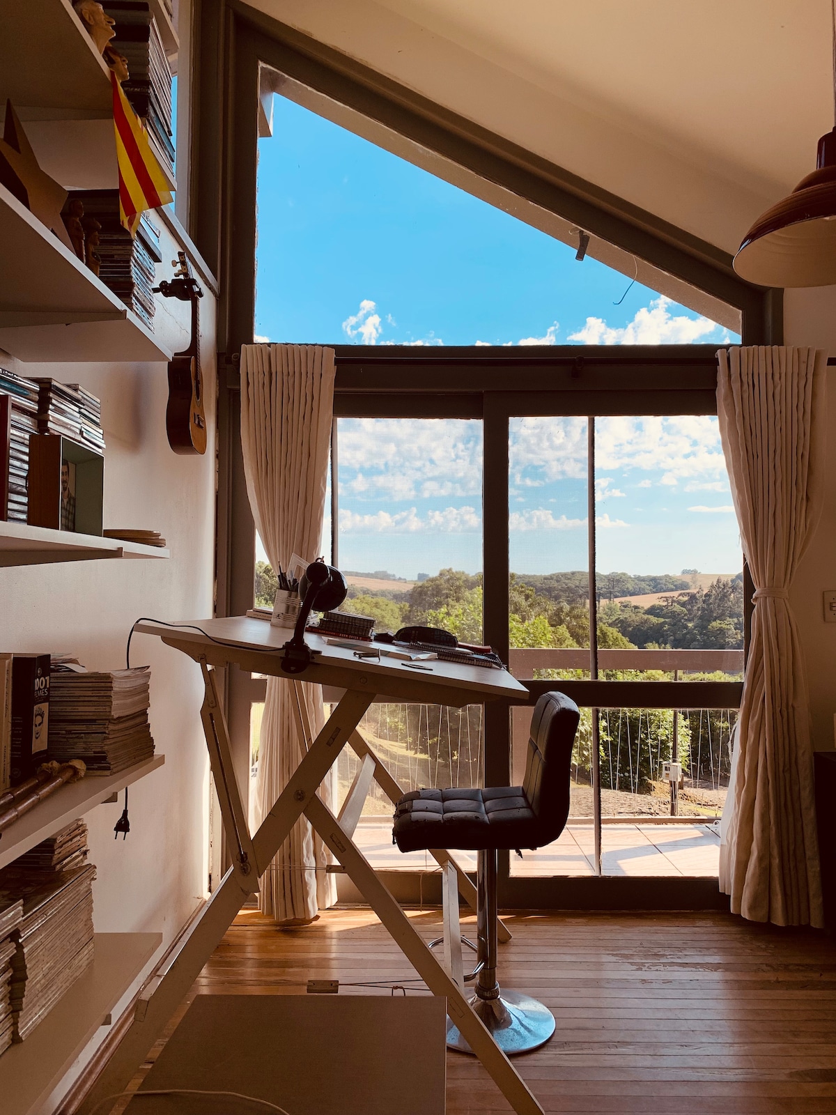 A bright workspace is positioned beside a large window, offering a panoramic view of the landscape. Natural light fills the room, illuminating bookshelves lined with books and a small collection of instruments. A modern, high-backed stool complements the desk.