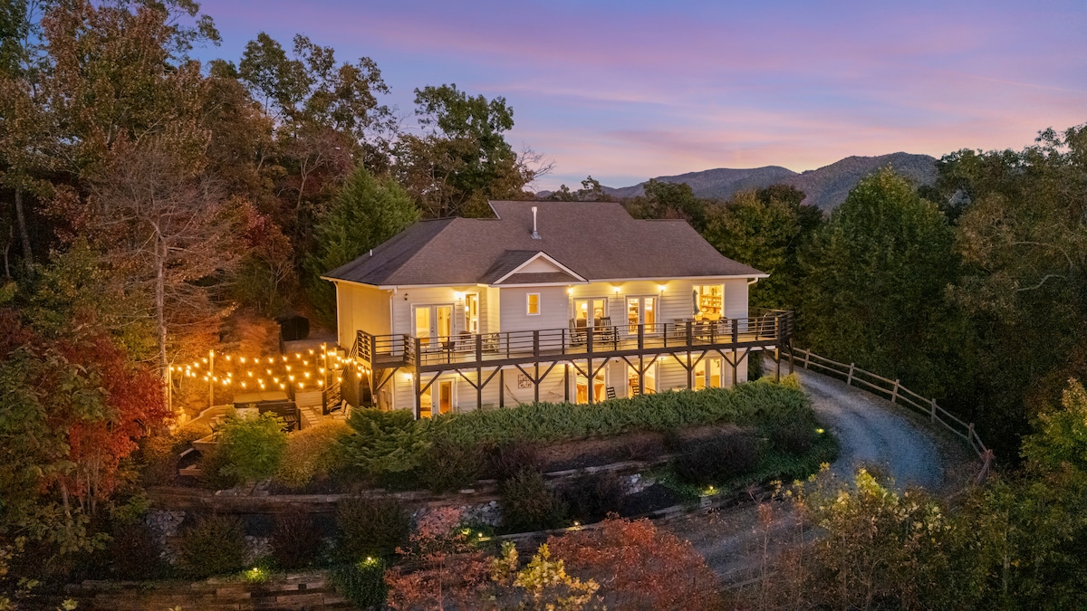 An aerial view of a spacious house surrounded by lush trees, showcasing soft evening lighting with string lights illuminating the deck. The scenic backdrop features mountains, creating a serene atmosphere enhanced by the natural landscape.