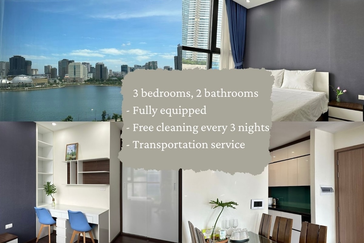 The image displays a bright and airy apartment interior with a large window showcasing a city view. A neatly made spacious bed is visible in one corner, while a dining area and a fully equipped kitchen are shown in another section. Modern furnishings and a calming color palette enhance the space.