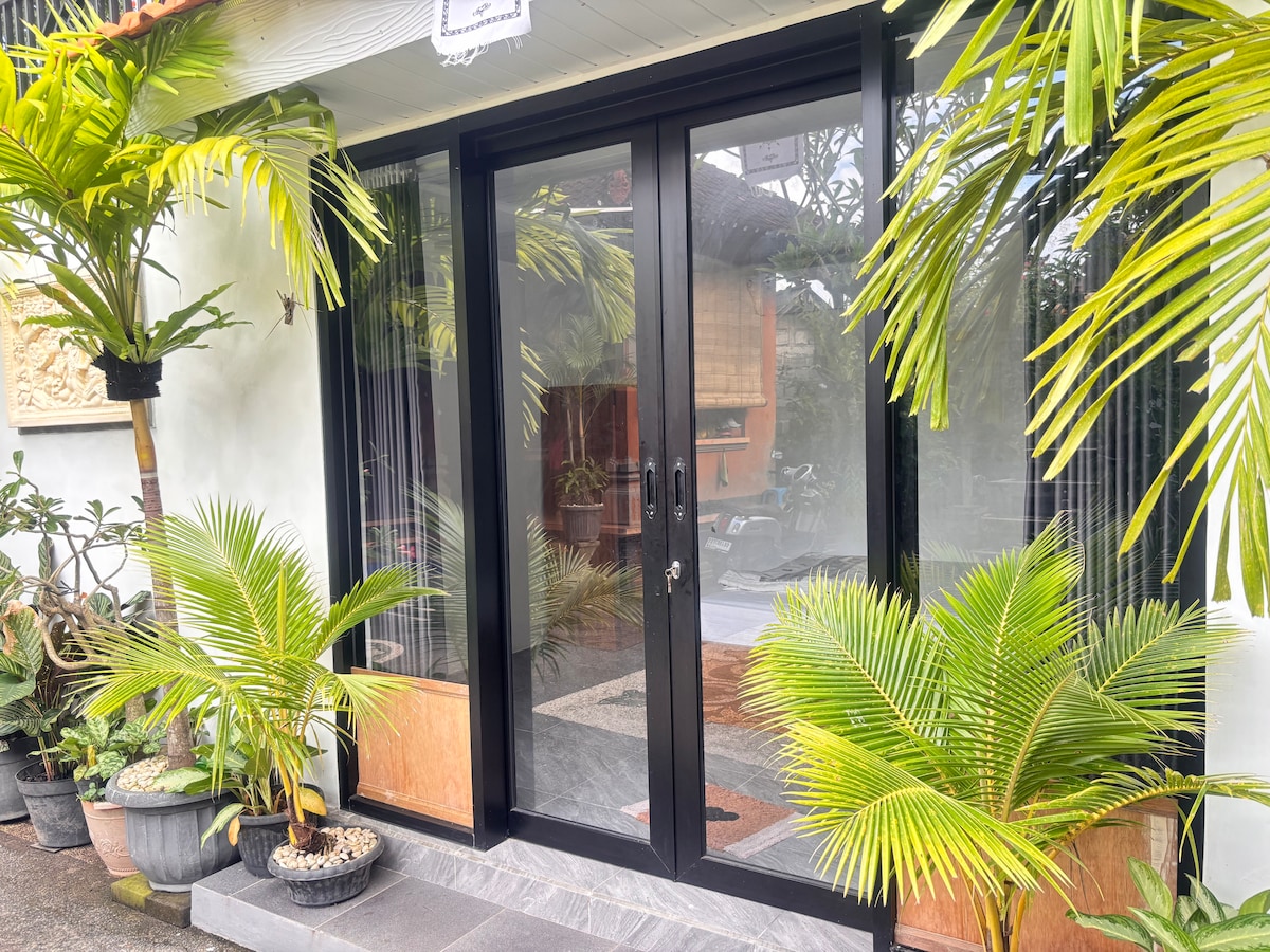 A modern entrance is flanked by lush green plants, providing a refreshing touch. The double glass doors, framed in black, invite natural light into the space. The contrasting stone flooring enhances the welcoming aspect of the entrance.