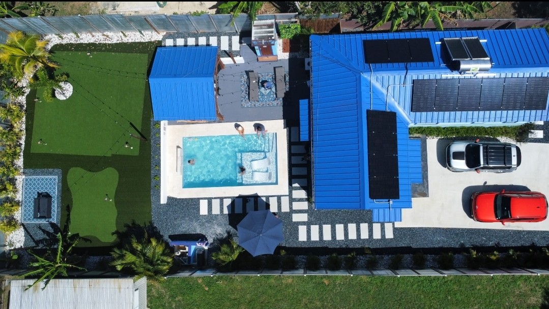 An aerial view captures the vibrant blue cottage with solar panels on the roof, surrounded by a landscaped yard. A private pool is visible alongside a mini-golf area, while several cars are parked in the driveway. Outdoor seating is arranged under an umbrella.