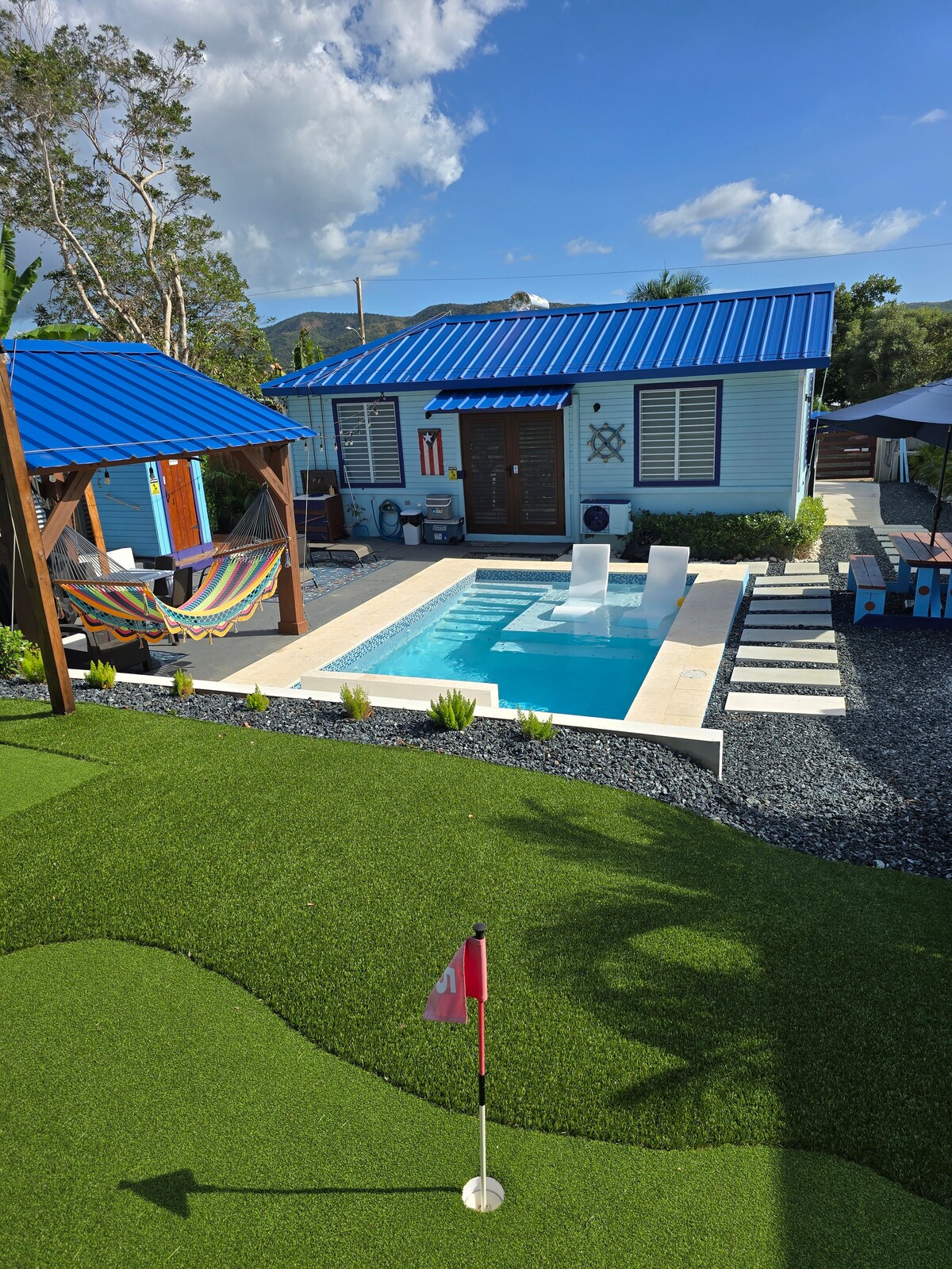 A private pool area is showcased with a turquoise pool surrounded by a well-maintained landscape. Green grass outlines the mini golf course, with white lounge chairs at the pool's edge. A colorful hammock is seen nearby, with the cottage in the background, topped by a bright blue roof.