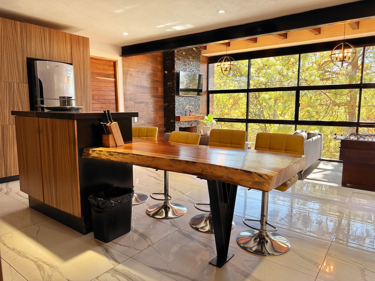 A modern dining area features a striking wooden table supported by a sleek black base and surrounded by comfortable yellow chairs. Large windows provide ample natural light and scenic views of the outdoors, while an adjoining kitchen area offers essential amenities.
