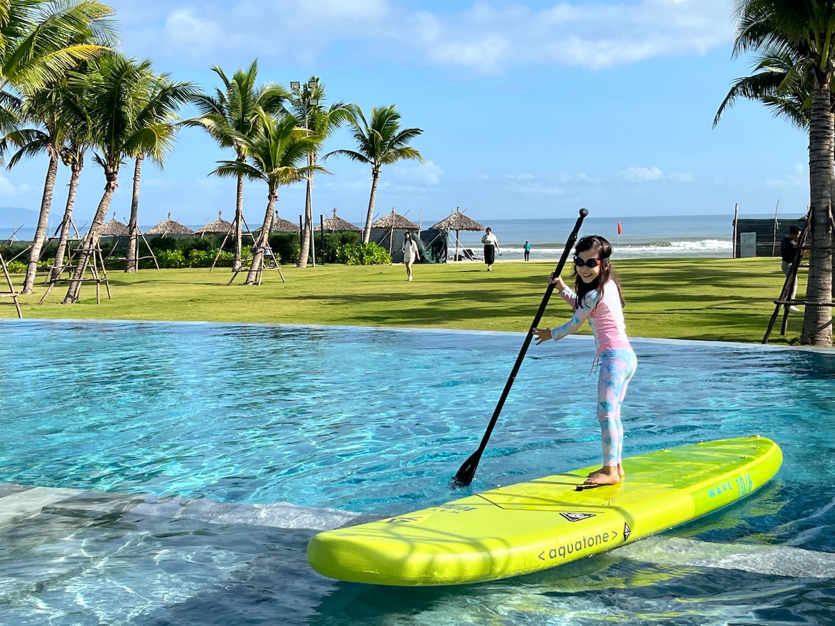 A child stands on a bright yellow paddleboard, navigating a calm pool with ocean views. Lush palm trees line the background, and beach huts are visible in the distance. Soft waves can be seen gently rolling onto the shore under a clear blue sky.