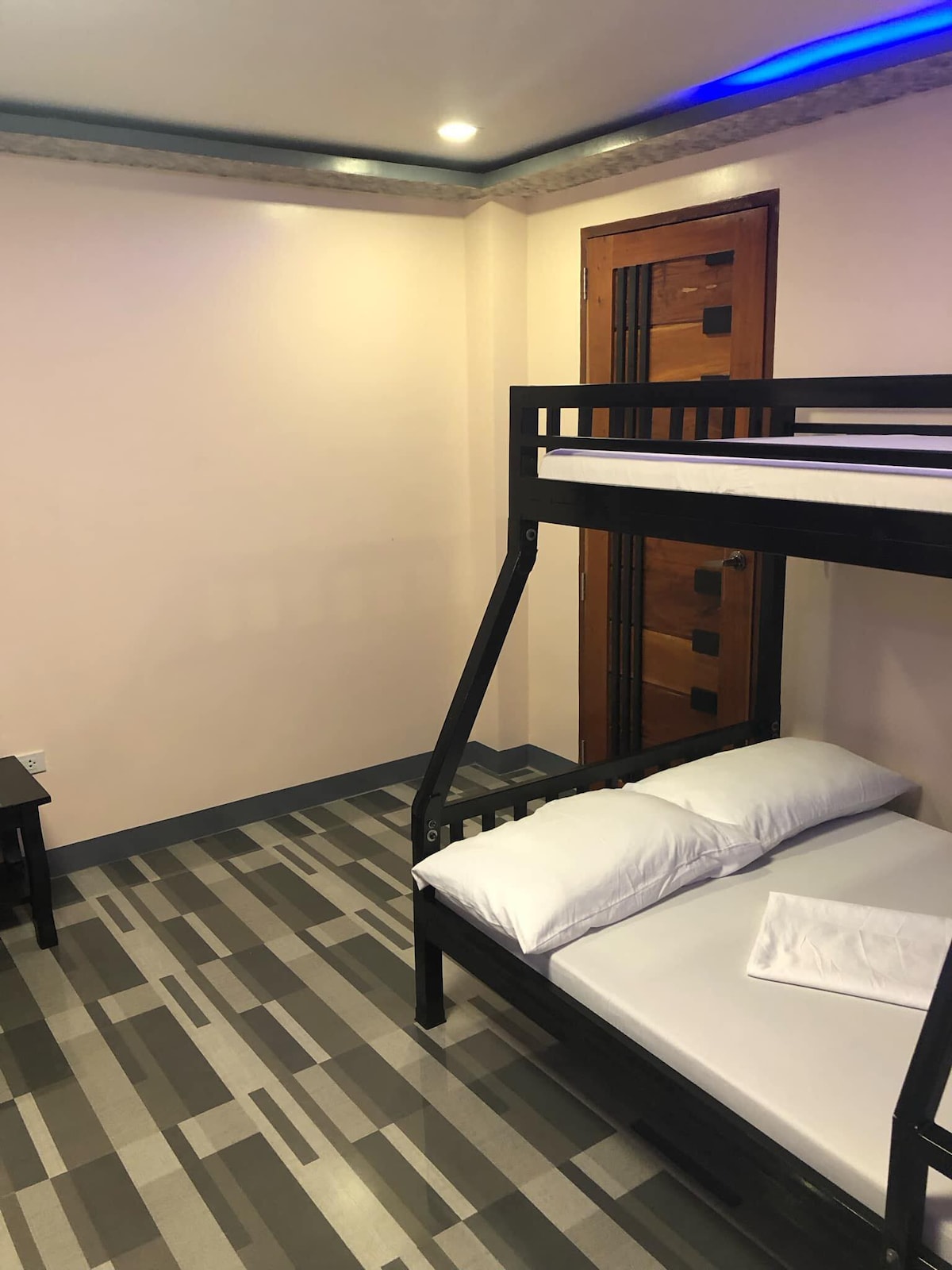 A minimalist room features a black wooden bunk bed with white mattresses and fresh linens. A small side table is positioned beside the bunk, and a wooden door with a detailed design complements the room's neutral walls and modern flooring.