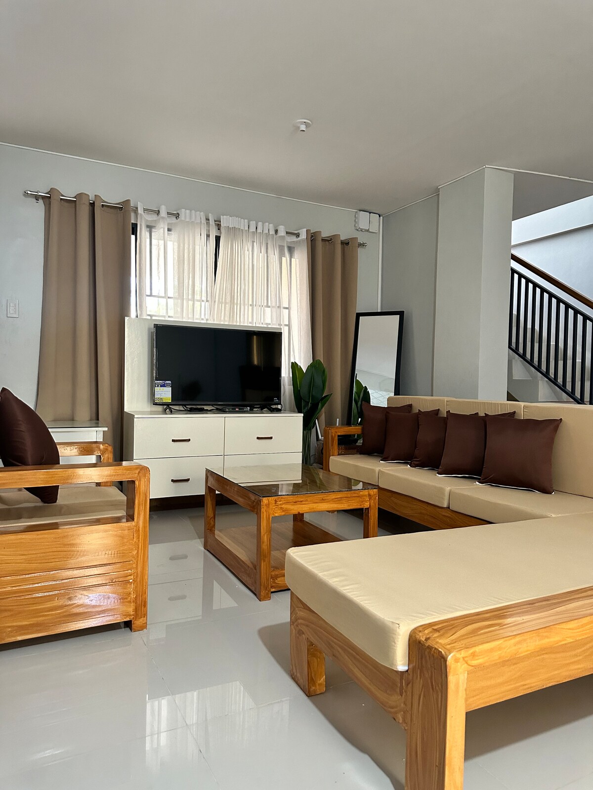 A modern living area showcases a combination of light-colored furnishings and natural wood accents. A large flat-screen television is positioned against a light wall, with a stylish mirror and potted plants included. Brown cushions adorn the sofa, enhancing the relaxed atmosphere.