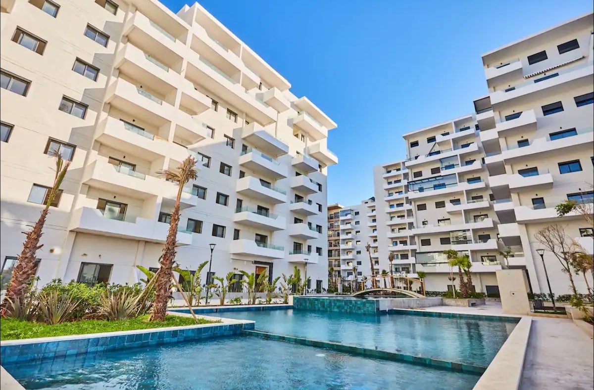 A modern apartment complex is showcased, featuring multiple white structures with balconies. A clear, inviting pool is situated in the foreground, surrounded by landscaped greenery and palm trees. Sunlight brightens the blue water, creating a refreshing atmosphere.