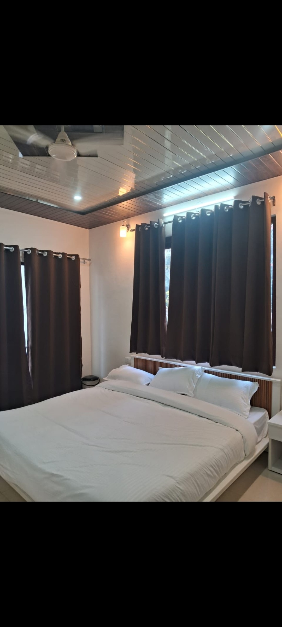 A bright sleeping area is furnished with a spacious bed covered with white linens. Dark curtains frame the windows, while a light fixture illuminates the ceiling. A nightstand is positioned beside the bed, offering a functional accent to the room.