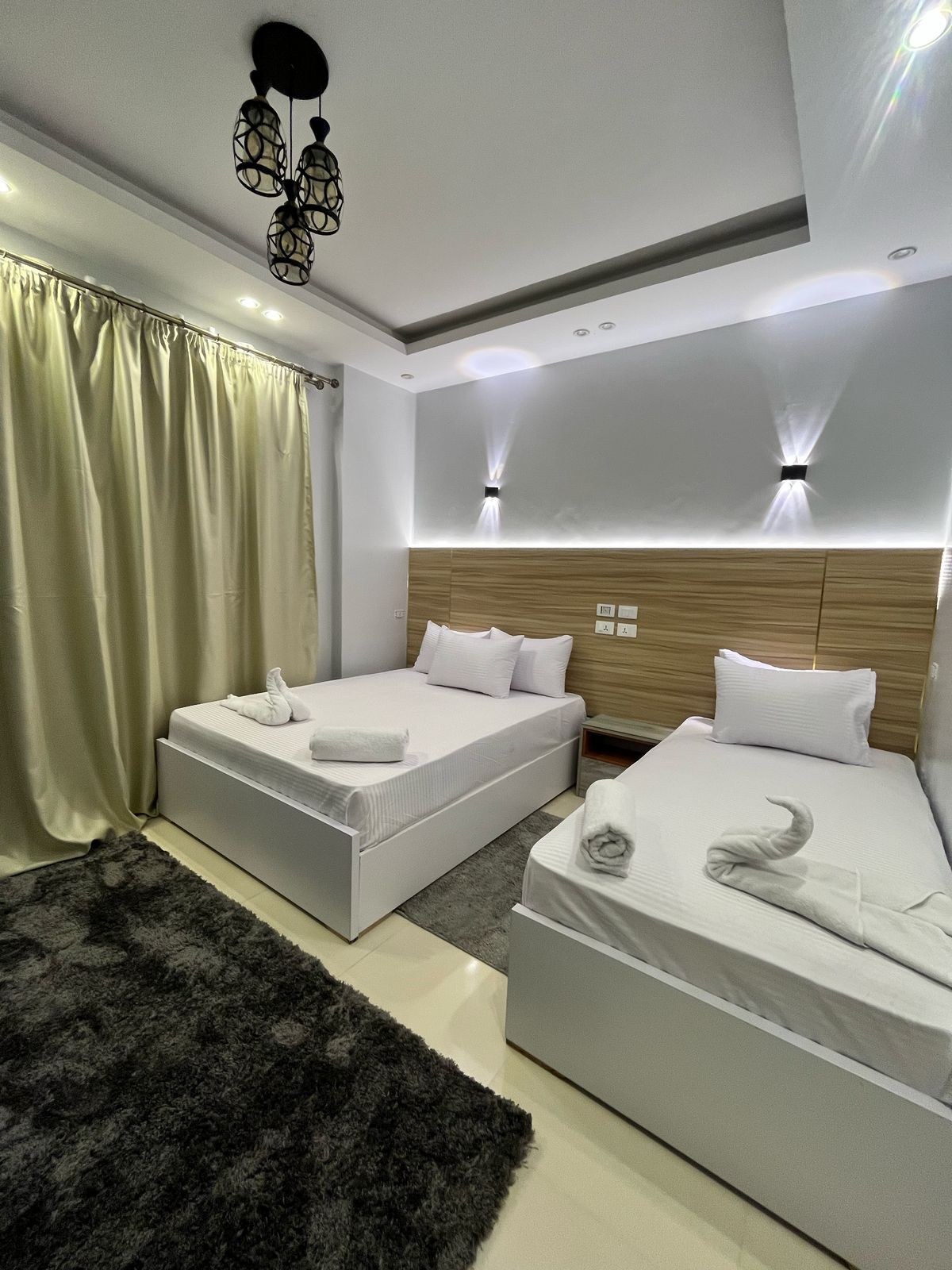 A well-lit sleeping area features two spacious beds, each dressed in crisp white linens. White bath towels are neatly arranged on the beds. Soft, neutral-toned curtains frame a window, while a plush gray rug covers the floor, contributing to a serene atmosphere.