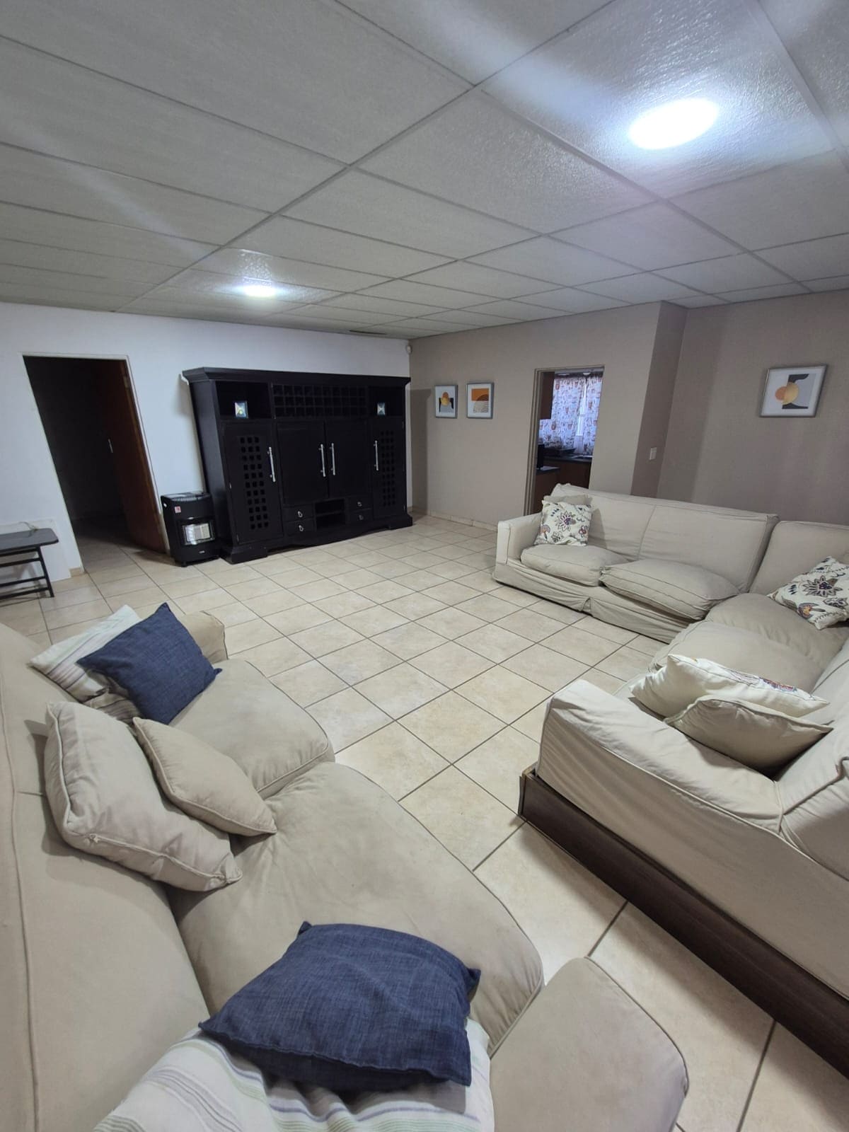 A spacious living area features multiple soft couches arranged to create a welcoming atmosphere. Neutral-toned tiles cover the floor, while walls are adorned with framed artwork. A large entertainment unit stands against one wall, complementing the overall design of the space.