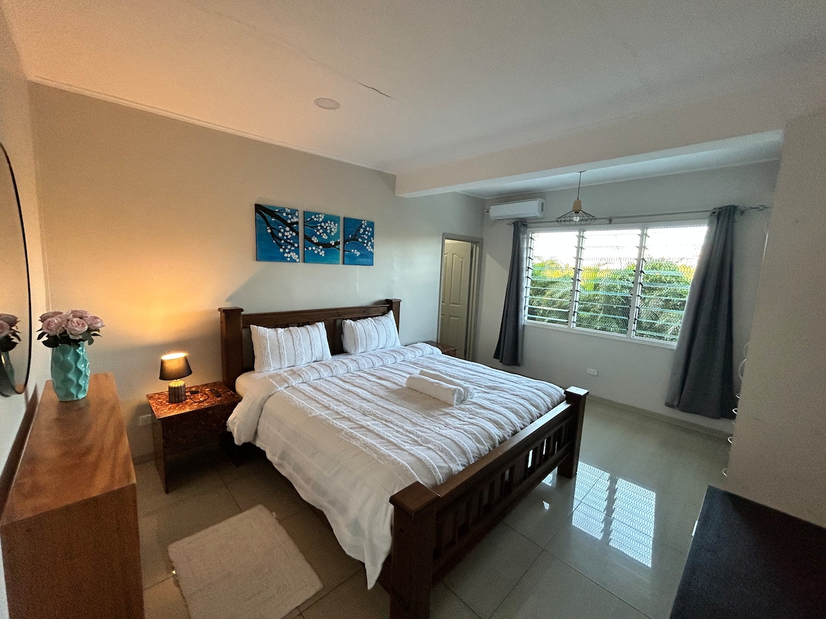 A spacious bedroom featuring a large wooden bed with ample bedding and a decorative arrangement of three blue artworks above. A bedside table with a lamp provides soft lighting, while a window allows natural light to enter. Light grey curtains frame the window, complementing the room's neutral palette.