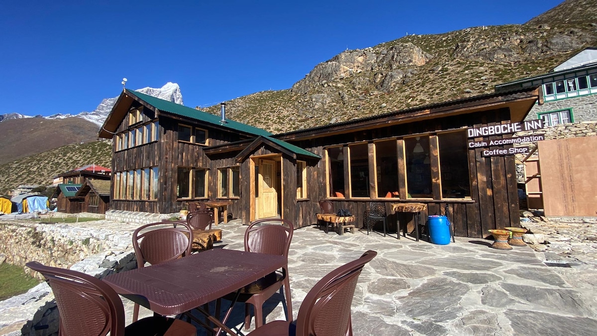 A rustic building featuring wooden architecture, with large windows that allow natural light to fill the interior. A stony patio is visible, furnished with several outdoor chairs and tables. Scenery of surrounding mountains adds to the peaceful environment.