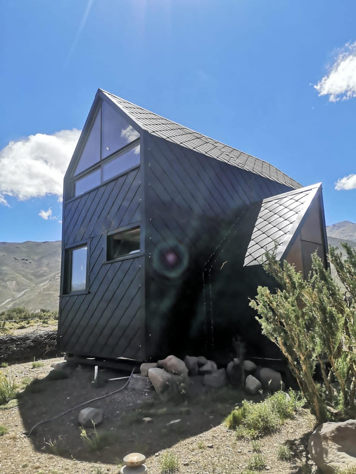 A modern black house is depicted against a clear blue sky, featuring distinct angular architecture. Large windows allow ample natural light, while nearby boulders and sparse vegetation highlight the surrounding landscape, creating a harmonious blend with the environment.