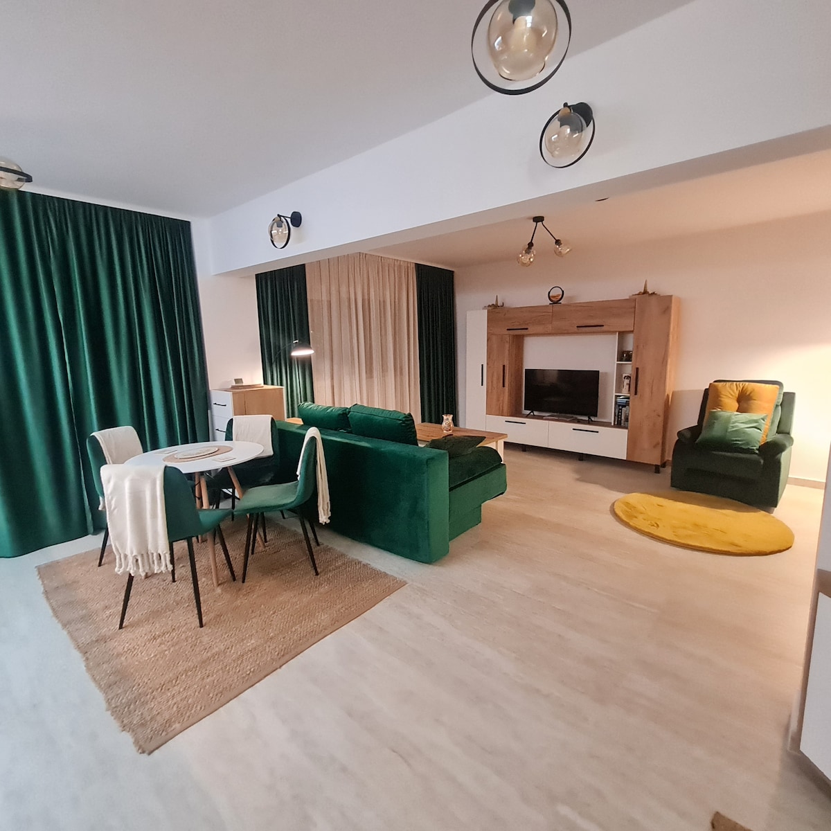 The living area is furnished with a green sofa and two armchairs, complemented by a circular yellow rug. A round dining table with black chairs is positioned nearby. Floor-to-ceiling curtains in emerald green add a touch of elegance, with wooden accents enhancing the space.