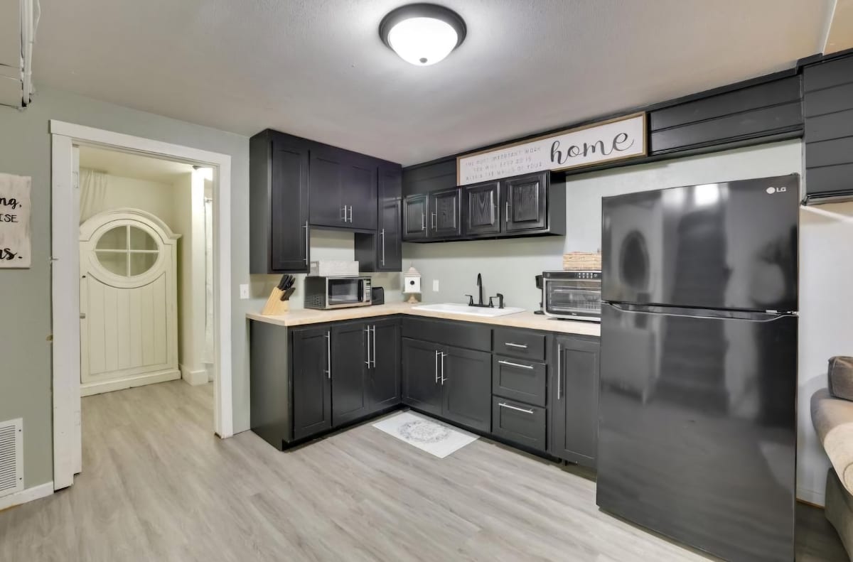 The kitchen features dark cabinetry and modern appliances, including a black refrigerator and two microwaves. A light fixture illuminates the space, which is complemented by a neutral-colored mat on the floor. A doorway leads to another area of the home.