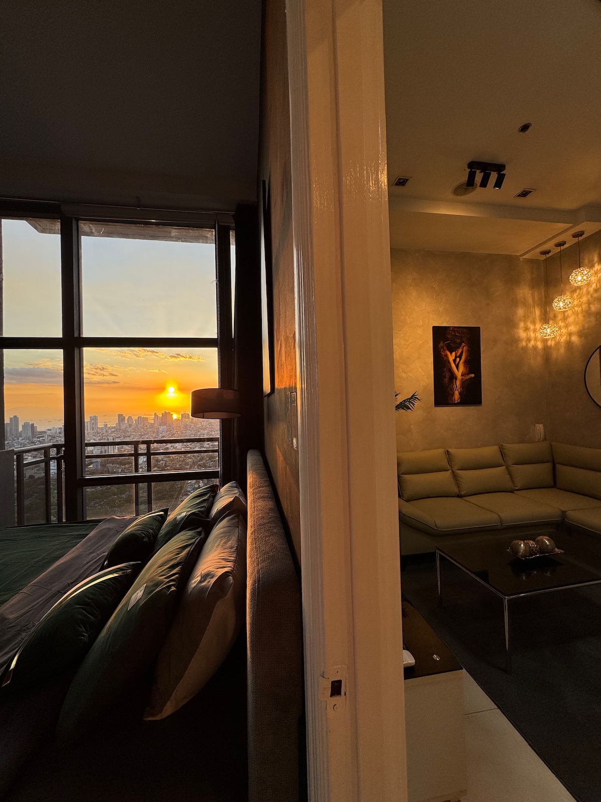 A spacious room features a king bed, with soft bedding visible. Sunlight pours in through large windows, illuminating the adjacent living area, which includes a modern couch and a coffee table. The city skyline and sunset can be seen through the glass, adding a warm ambiance.