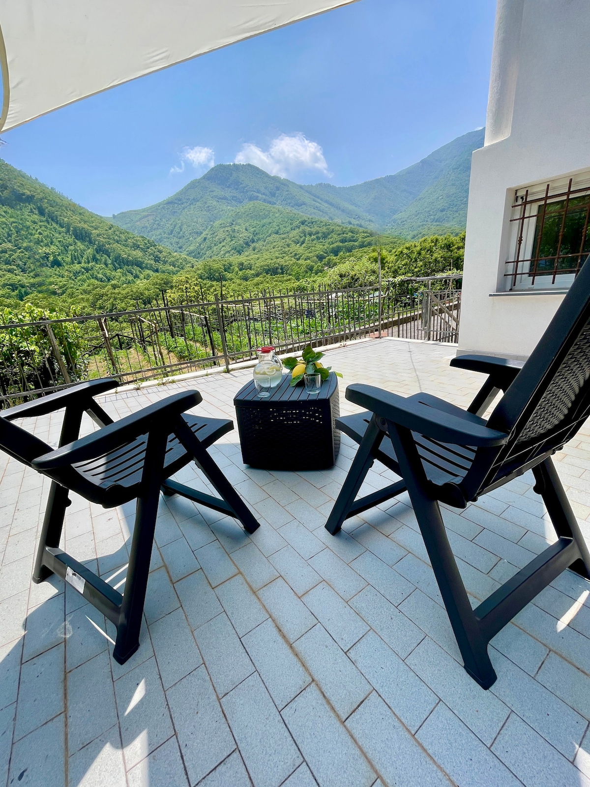 An outdoor seating area is presented, featuring two black chairs positioned alongside a small table. Lush green mountains are visible in the background, with the sky lit by daylight. A refreshing beverage, including fruits, is placed on the table, enhancing the serene atmosphere.