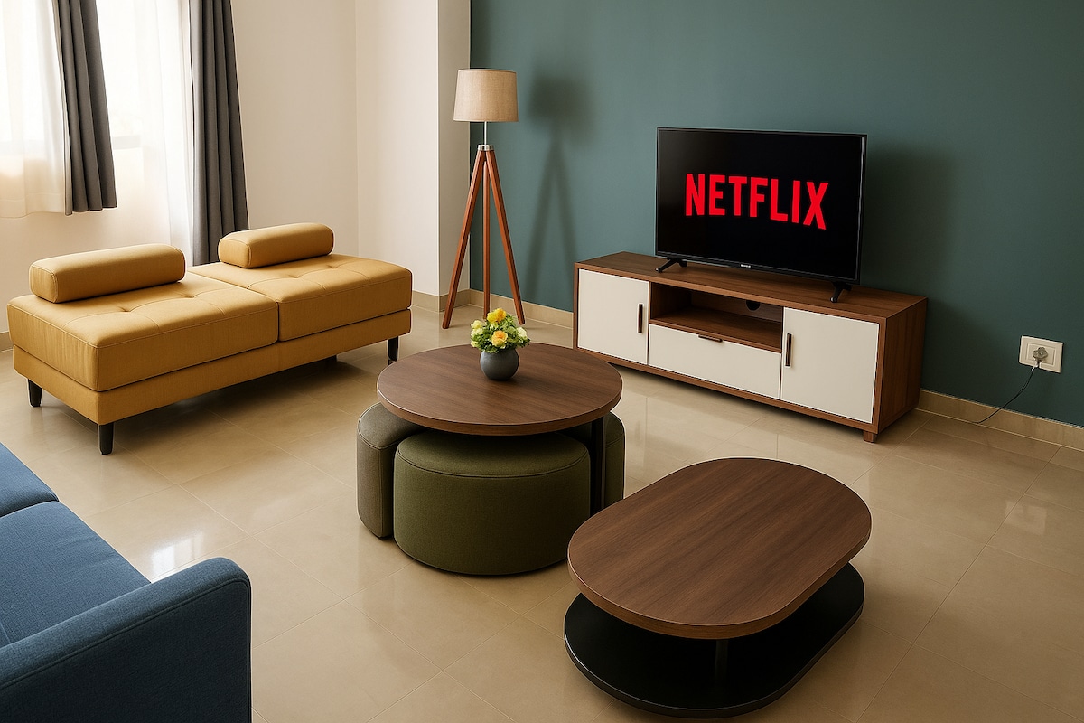 A modern living area is highlighted by a yellow sofa and a blue couch, paired with round coffee tables. A wooden entertainment unit hosts a television displaying the Netflix logo. Natural light streams through windows, illuminating the neutral-colored floor and the green accent wall.