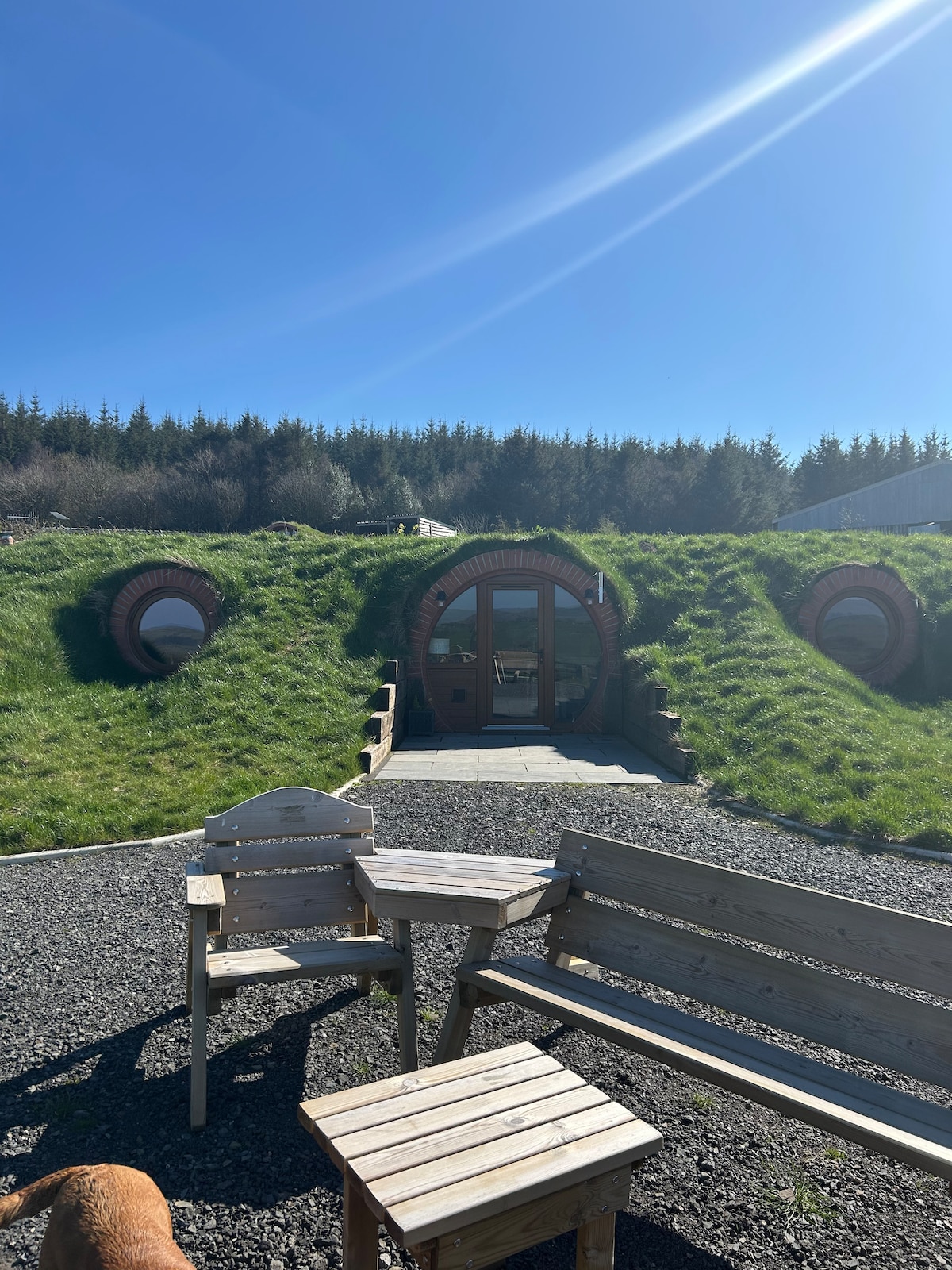 The image showcases a hobbit pod nestled within a grassy landscape, framed by natural surroundings. Two round windows are visible on the pod’s façade. In the foreground, wooden furniture elegantly complements the outdoor setting, inviting relaxation in the sunlit atmosphere.